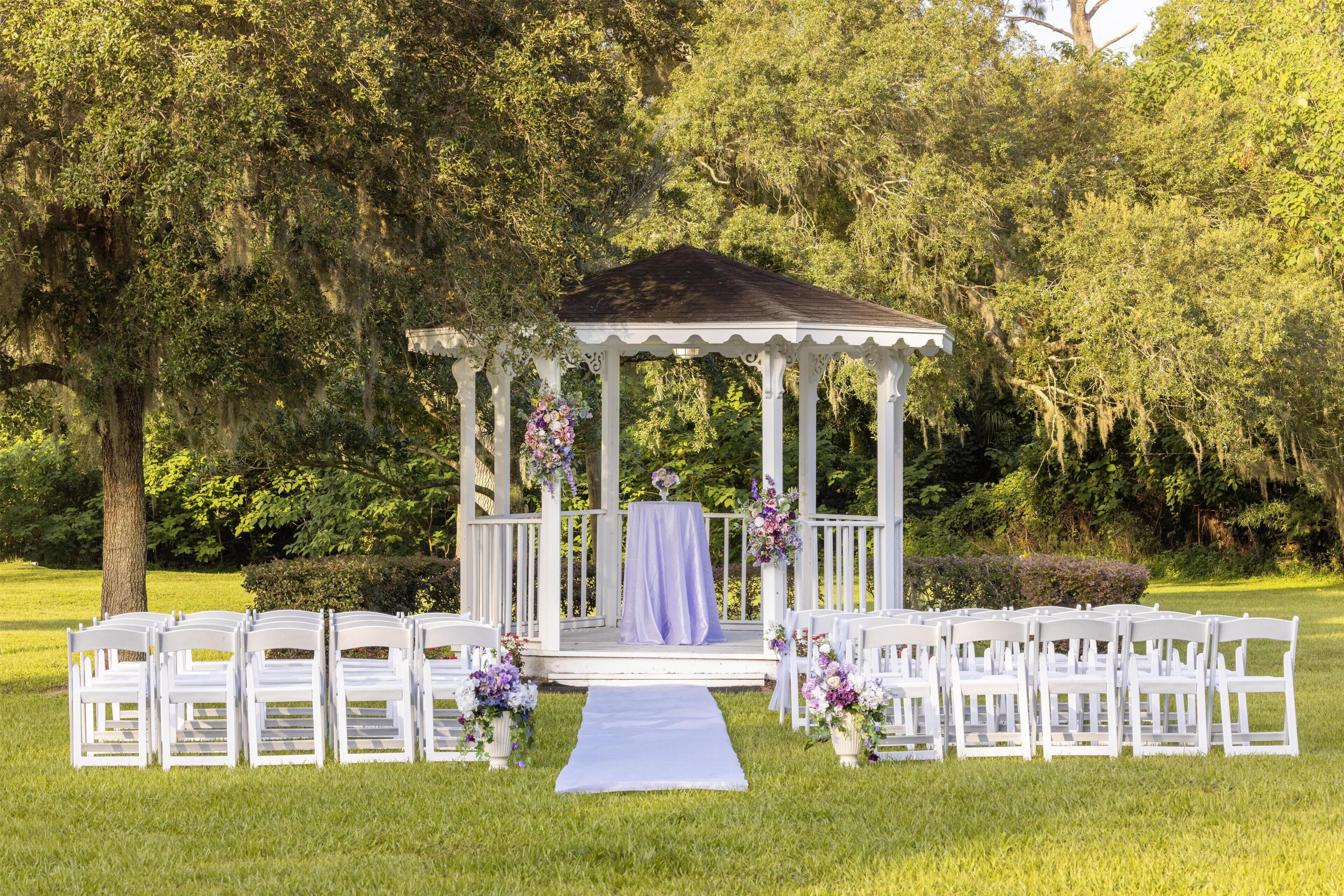 A gazebo with a white exterior and a purple tablecloth, surrounded by white chairs, set on a grassy lawn with trees in the background.