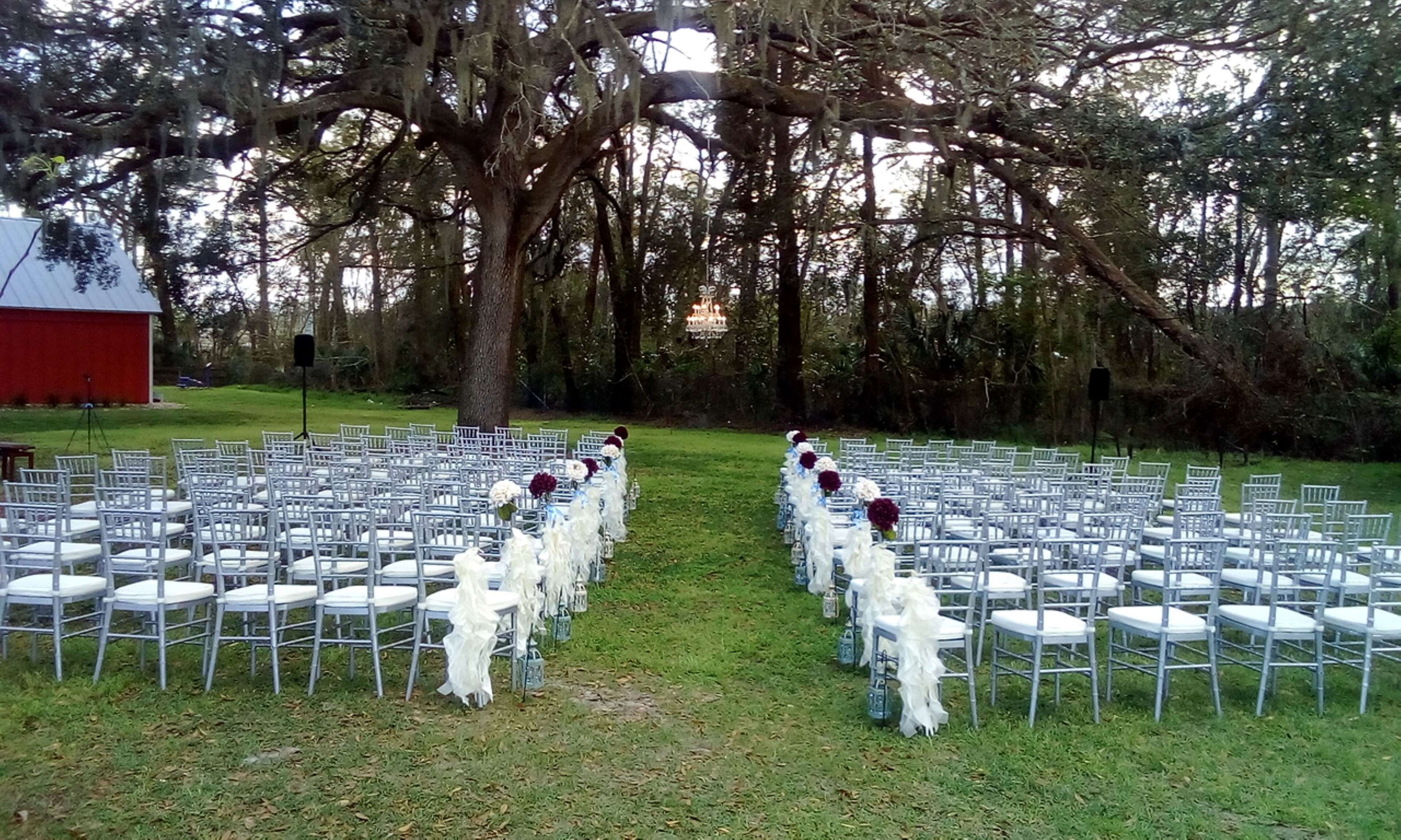 An outdoor wedding venue with rows of white chairs and tables, a large tree in the center, and a red building in the background.