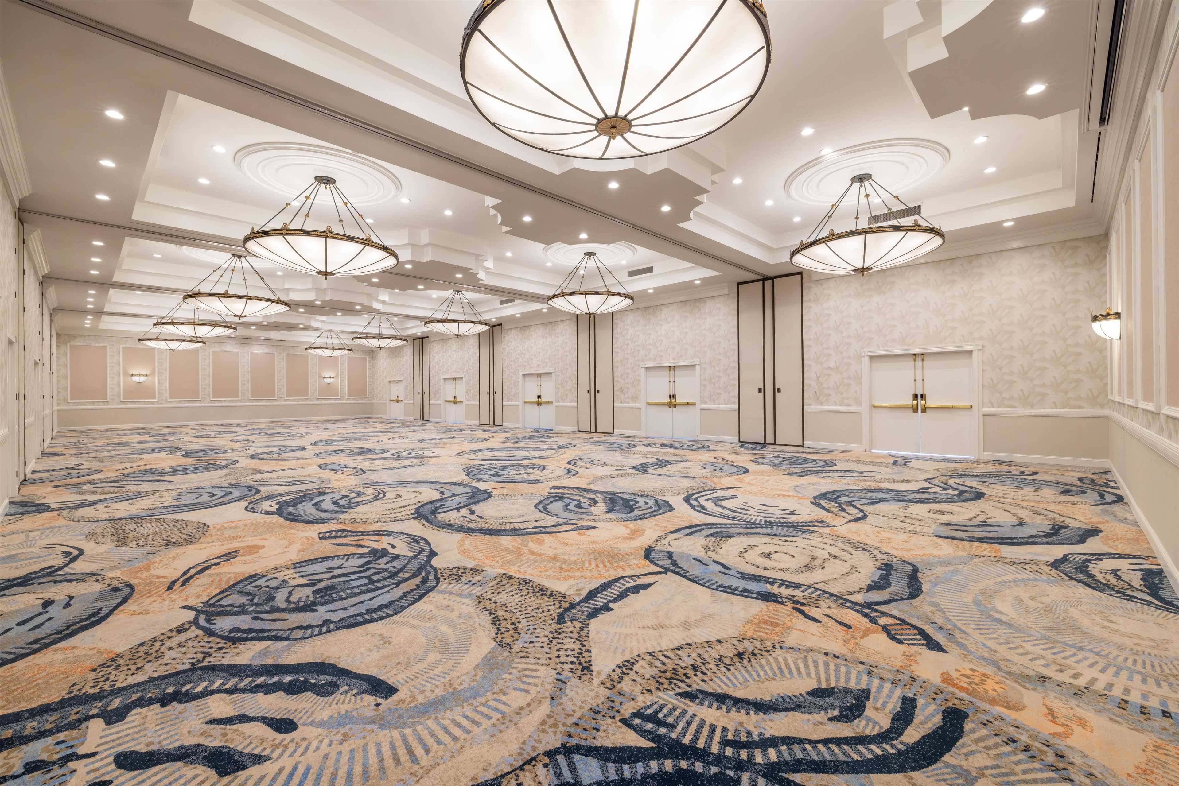 A spacious hotel lobby with a patterned carpet, multiple chandeliers, and a symmetrical room layout.