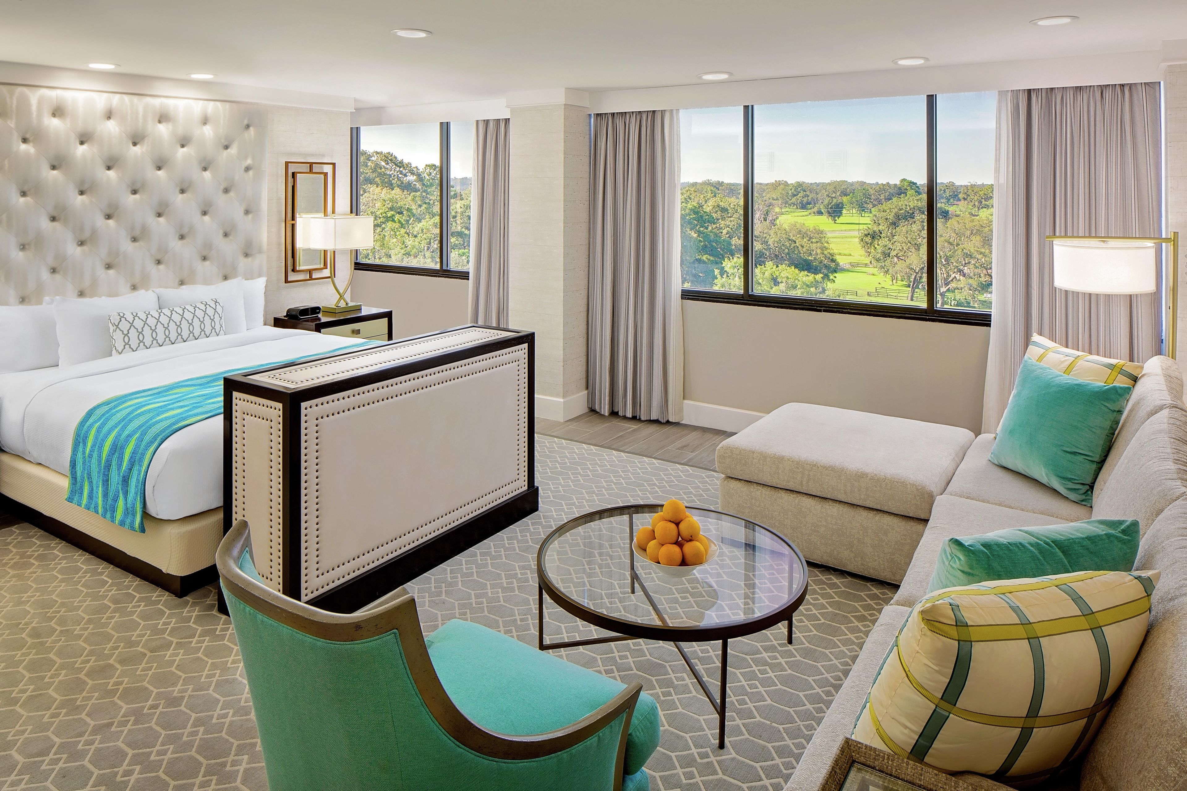 Una habitación de hotel con cama, sofá, sillón, una mesa con naranjas y ventanas amplias con vistas a la vegetación.