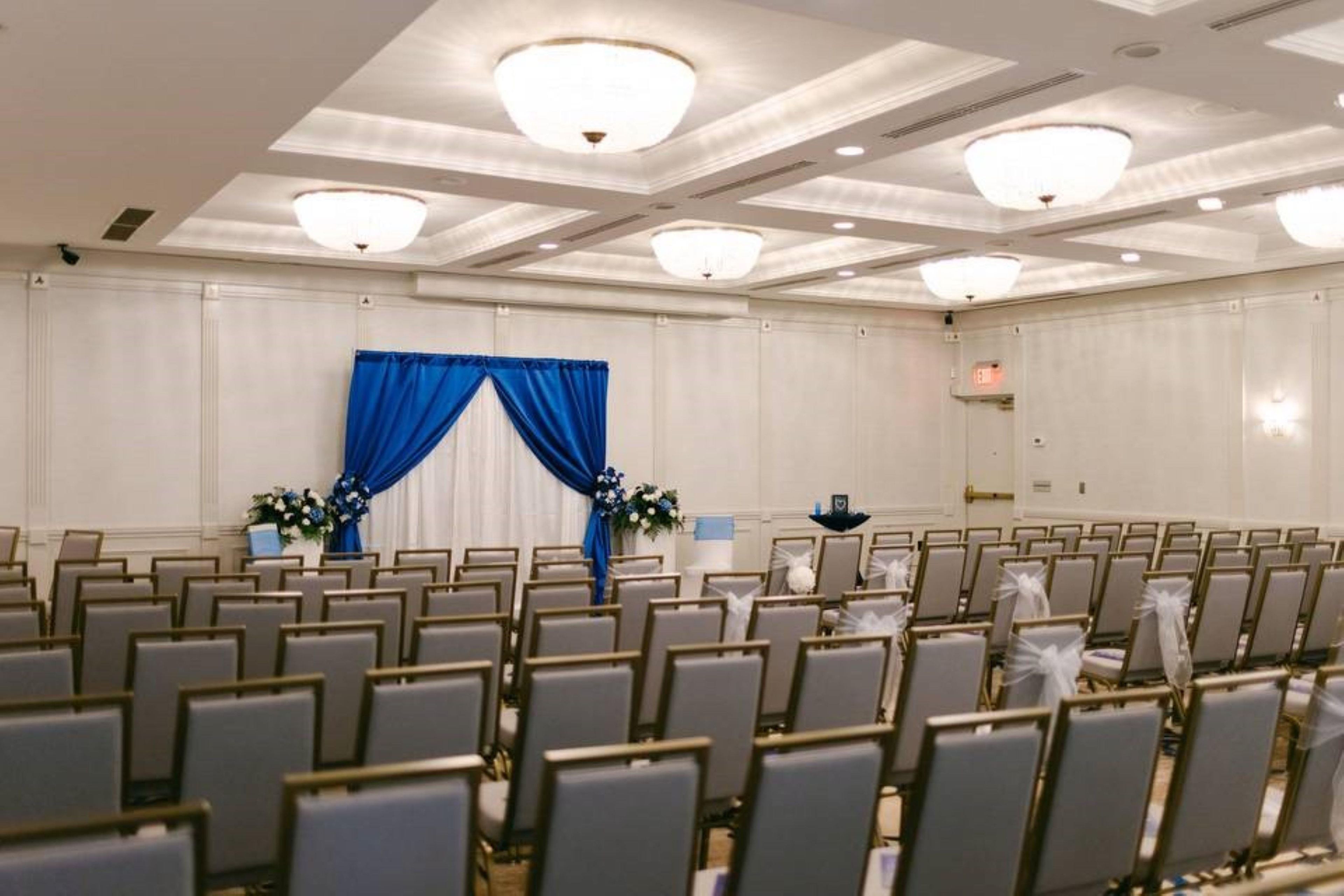 Un salón de conferencias con filas de sillas, un escenario con cortinas azules y luces en el techo.