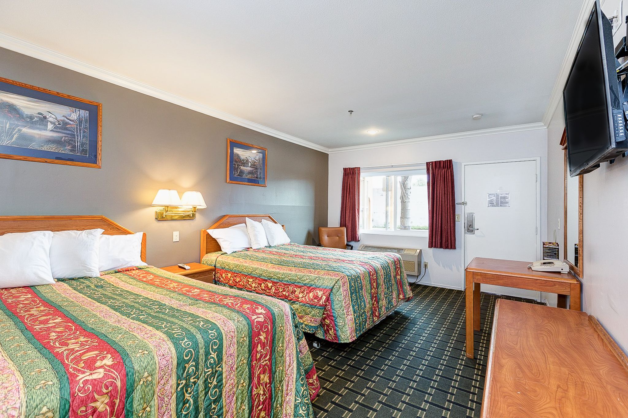 A hotel room with two beds, a TV, a desk, and a window with curtains.