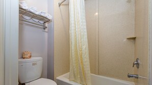 A bathroom with a toilet, a shower with a yellow curtain, and a shelf with folded towels. - Buena Vista Inn (Anaheim)