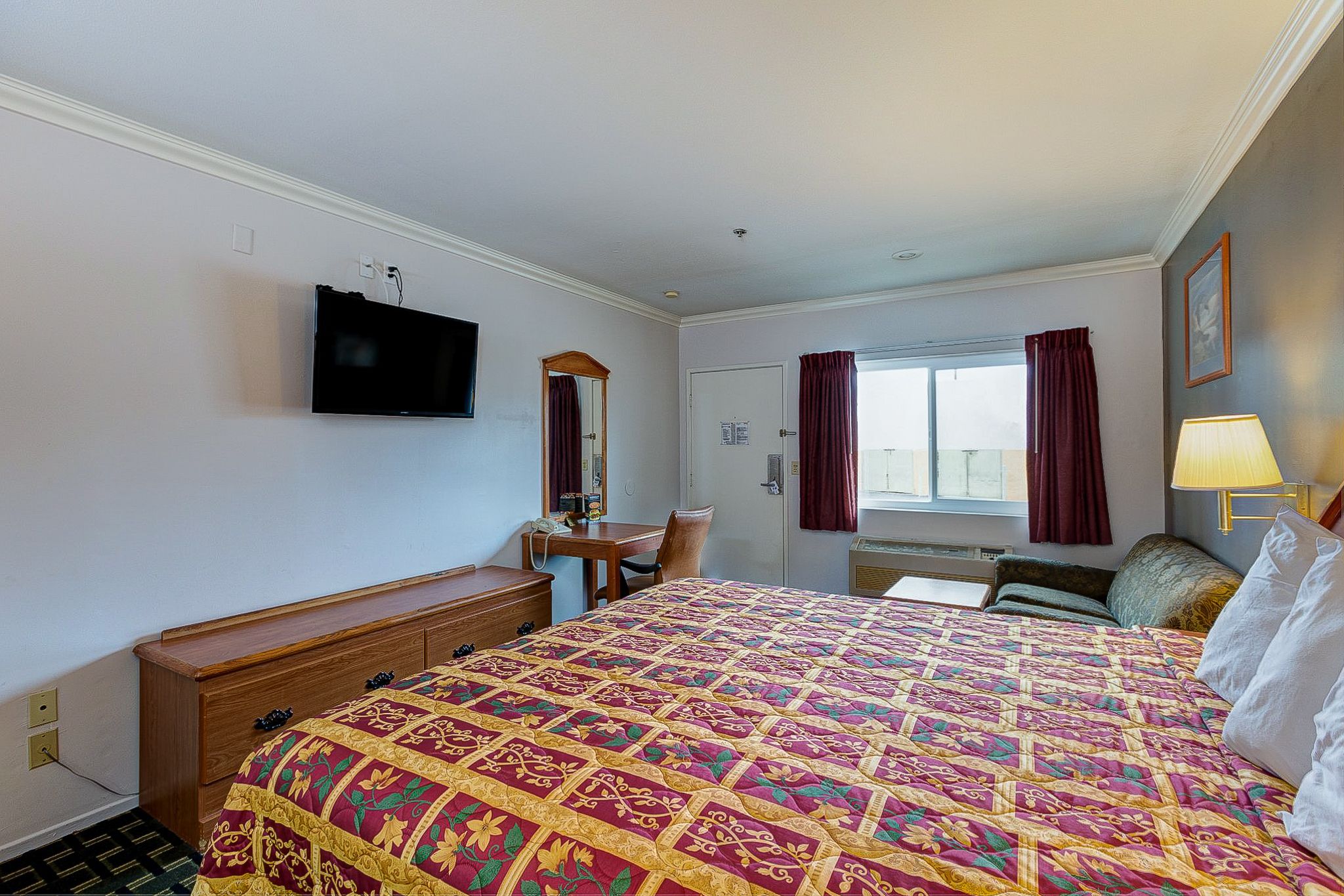Una habitación de hotel con una cama grande, un escritorio, una silla, un televisor montado en la pared y una ventana con cortinas.