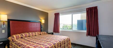A hotel room with a large bed, a desk, a window with curtains, and a lamp on the nightstand.