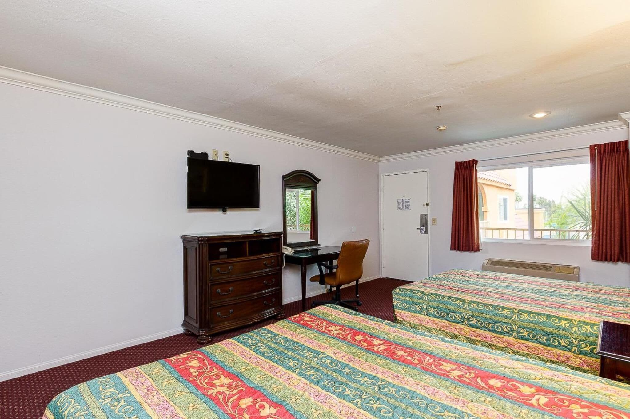 A hotel room with two beds, a desk, a dresser, a television, and a window with curtains.