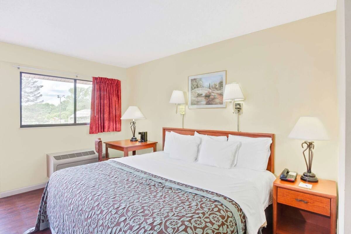 A hotel room with a bed, two bedside tables, a desk, a window with curtains, and a painting on the wall.