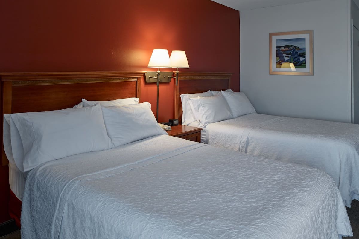 Two single beds with white linens, a wooden headboard, a nightstand with a lamp, and a framed picture on the wall.