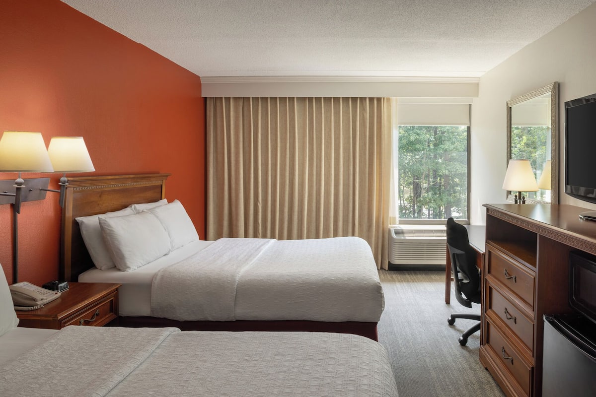 A hotel room with two beds, a desk, a dresser, a TV, and a window with curtains.