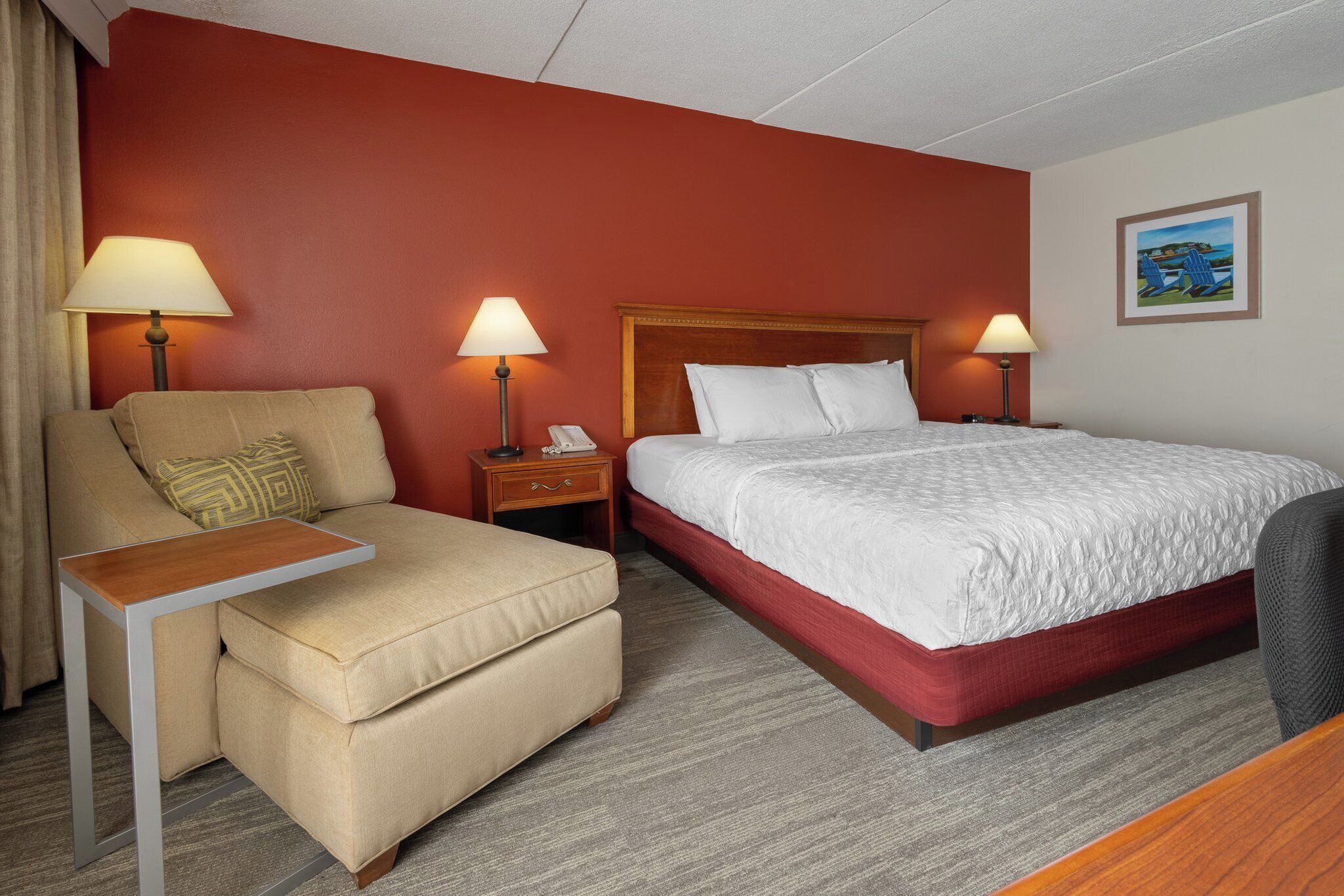 A hotel room with a bed, a desk, a chair, a nightstand, and a lamp.