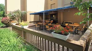 An outdoor patio with wooden tables and chairs, covered by a blue awning, and surrounded by potted plants and flowers. - Long Creek Hotel by IHG (South Portland)