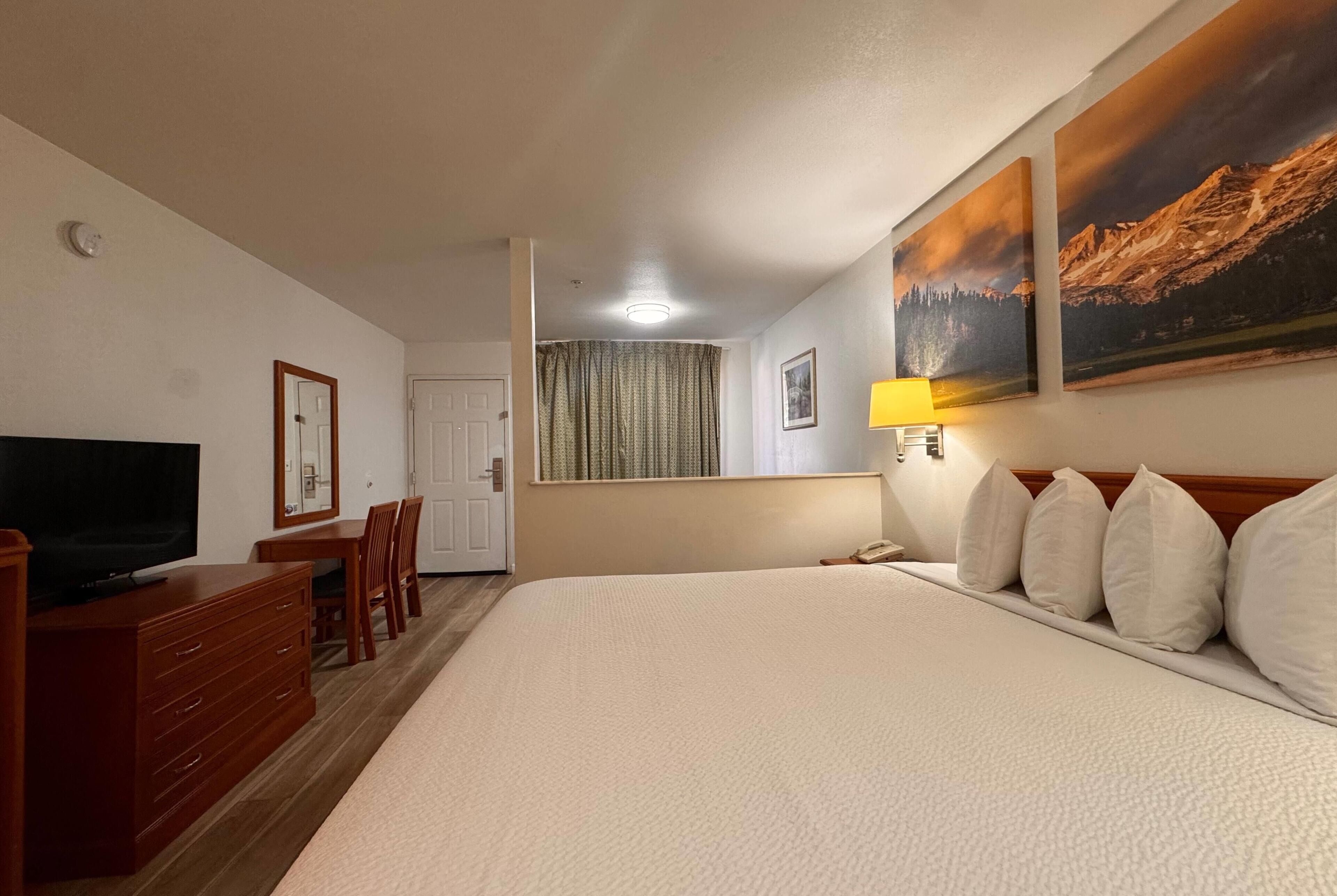 Une chambre d’hôtel comprenant un lit, une télévision, un bureau avec des chaises, une lampe et deux tableaux accrochés au mur.