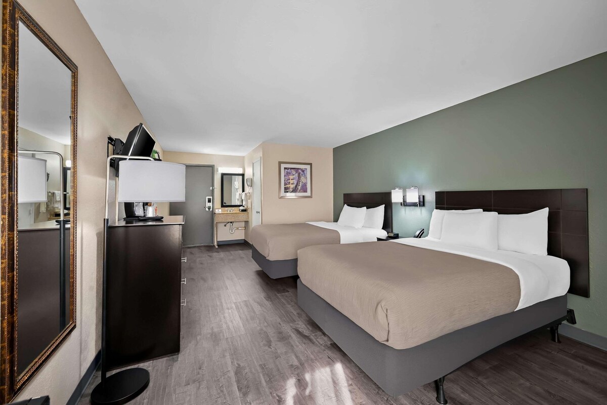 A hotel room with two beds, a dresser, a mirror, a desk with a lamp, and a framed picture on the wall.