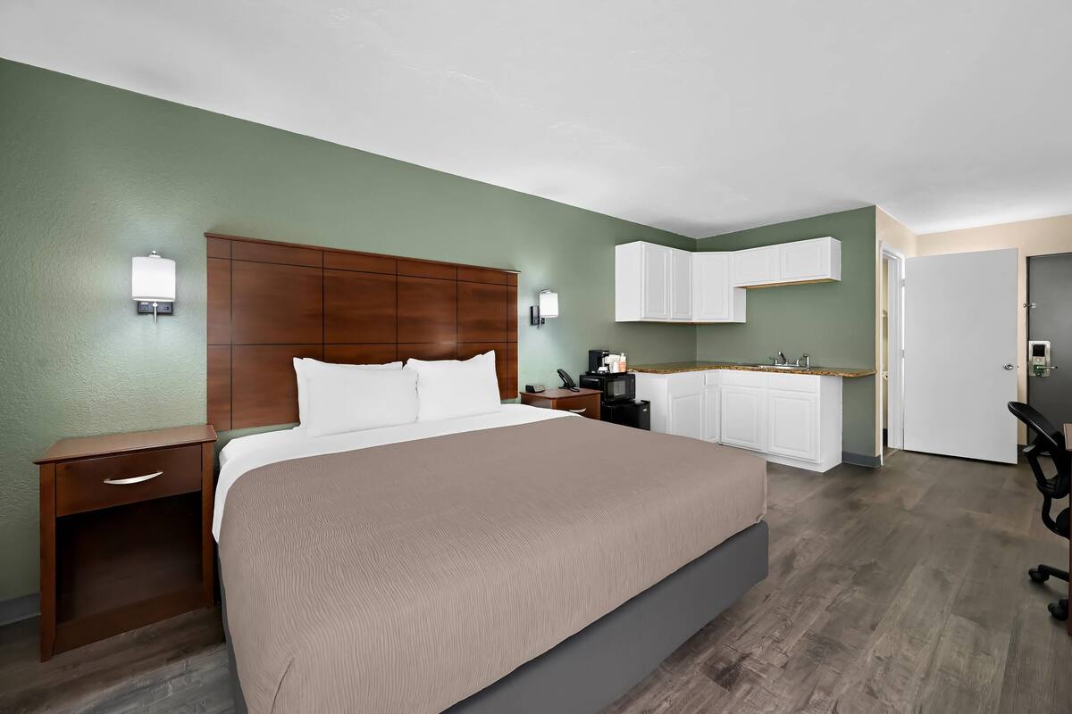 Habitación de hotel con una cama grande, una mesita de noche de madera, un escritorio con una silla, una kitchenette y un baño.