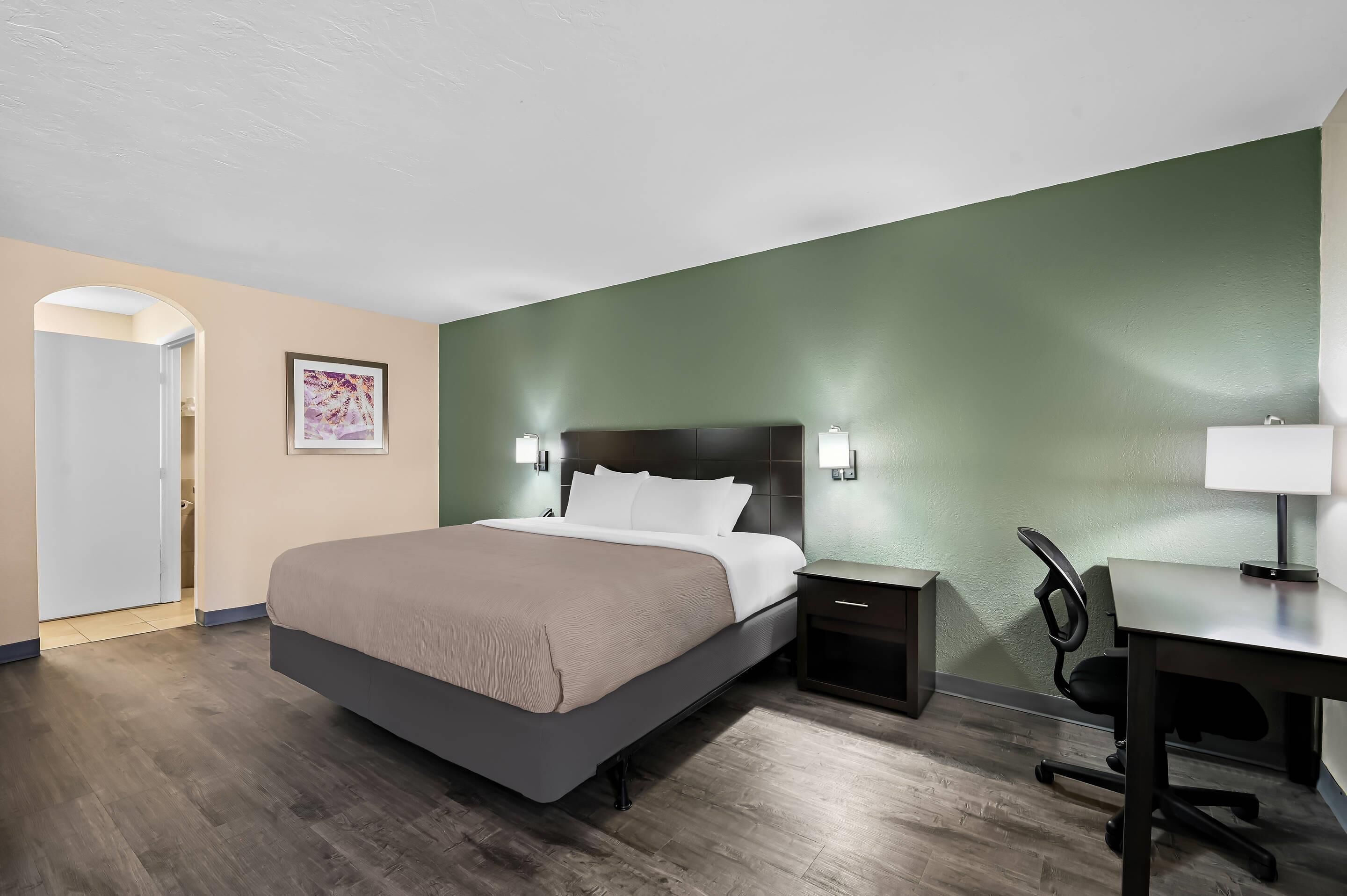 Une chambre d’hôtel avec un lit, une table de chevet, un bureau, une chaise et un tableau au mur.