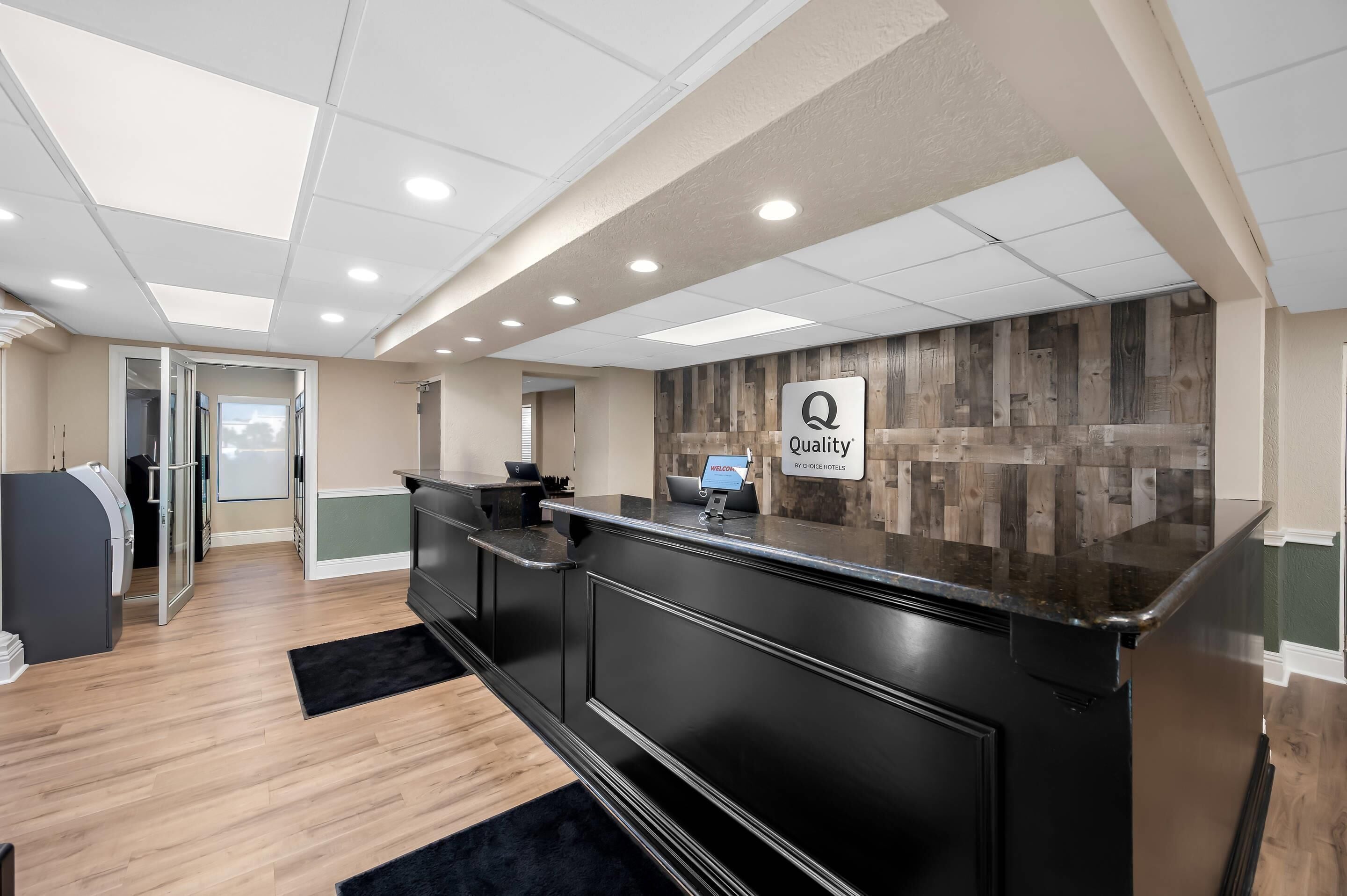 Un espace d’accueil d’hôtel au design moderne, comprenant un comptoir d’accueil, un écran numérique et un panneau indiquant « Qualité garantie par Oriole Hotels ».