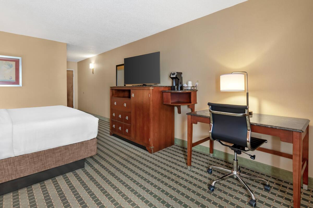 Habitación de hotel con cama, escritorio con silla, televisor, cafetera y lámpara.