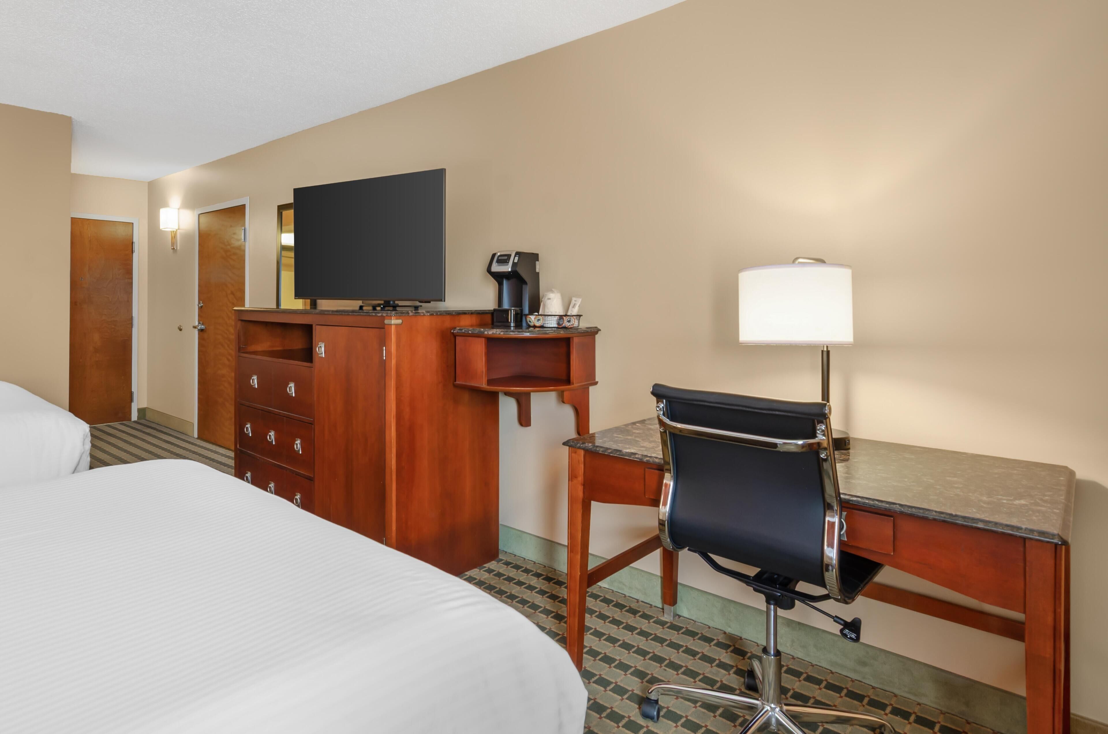 Quarto de hotel com cama, escrivaninha com cadeira, televisão, cafeteira e abajur.