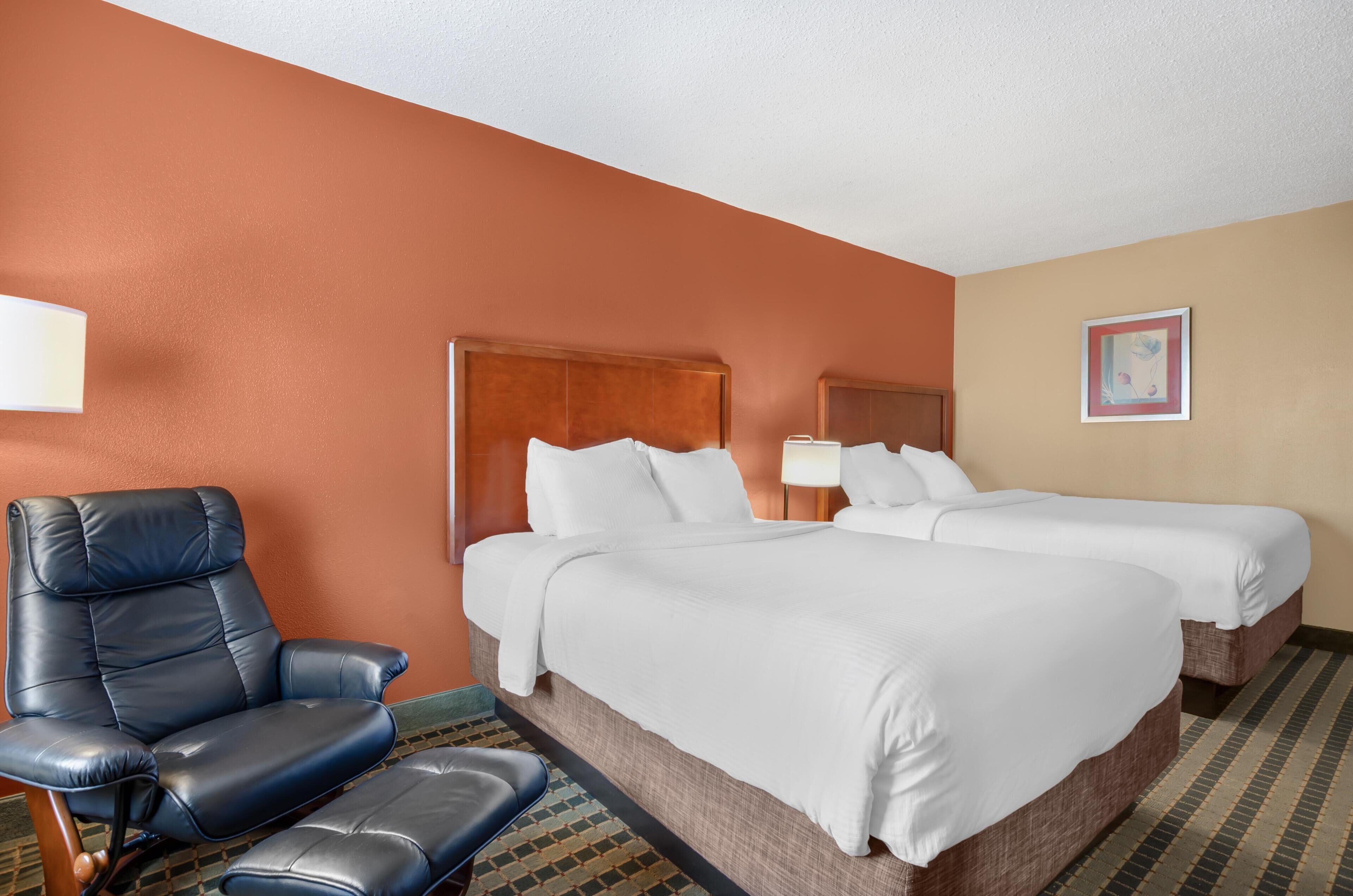 Quarto de hotel com duas camas, uma cadeira, um abajur e um quadro na parede.