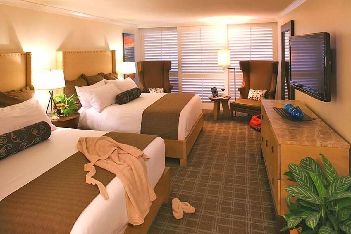 Ein Hotelzimmer mit zwei Betten, einem Schreibtisch mit Computer, einem Fernseher und einem großen Fenster mit Jalousien.