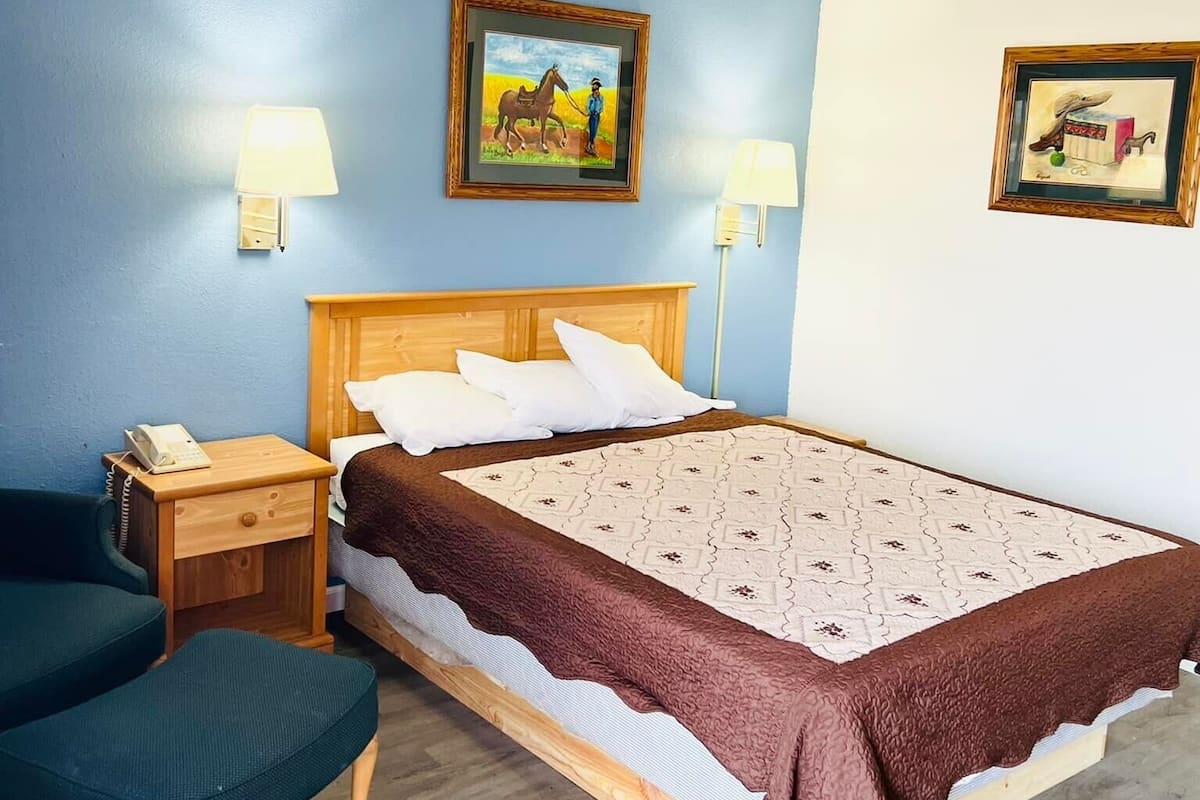 A bedroom with a wooden bed, two bedside lamps, a nightstand with a phone, a green chair, and two framed pictures on the wall.