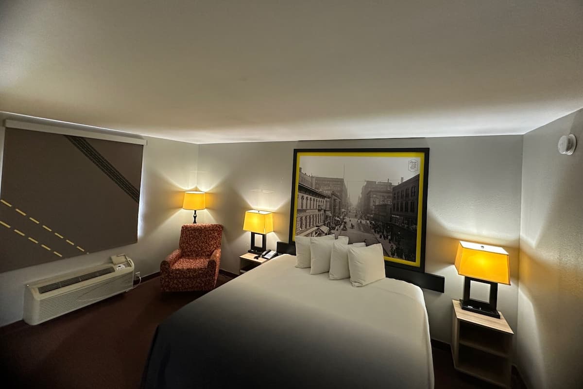 A hotel room with a large bed, two bedside lamps, a chair, a radiator, and a framed picture on the wall.