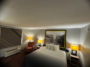 Habitación de hotel con una cama grande, dos lámparas de noche, una silla, un radiador y un cuadro en la pared.