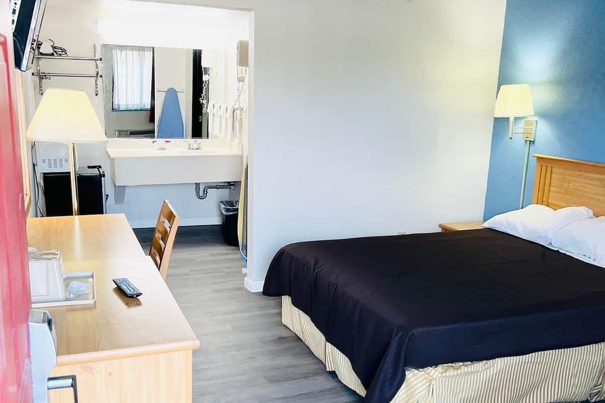 A hotel room with a bed, a desk with a chair, a mirror, a television, and a lamp.