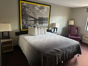 Habitación de hotel con una cama grande, dos mesitas de noche con lámparas, una silla y un cuadro grande con un puente sobre el agua.