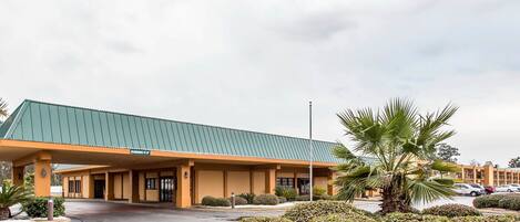 A hotel with a green roof and orange facade, featuring a parking lot and landscaped areas with palm trees.