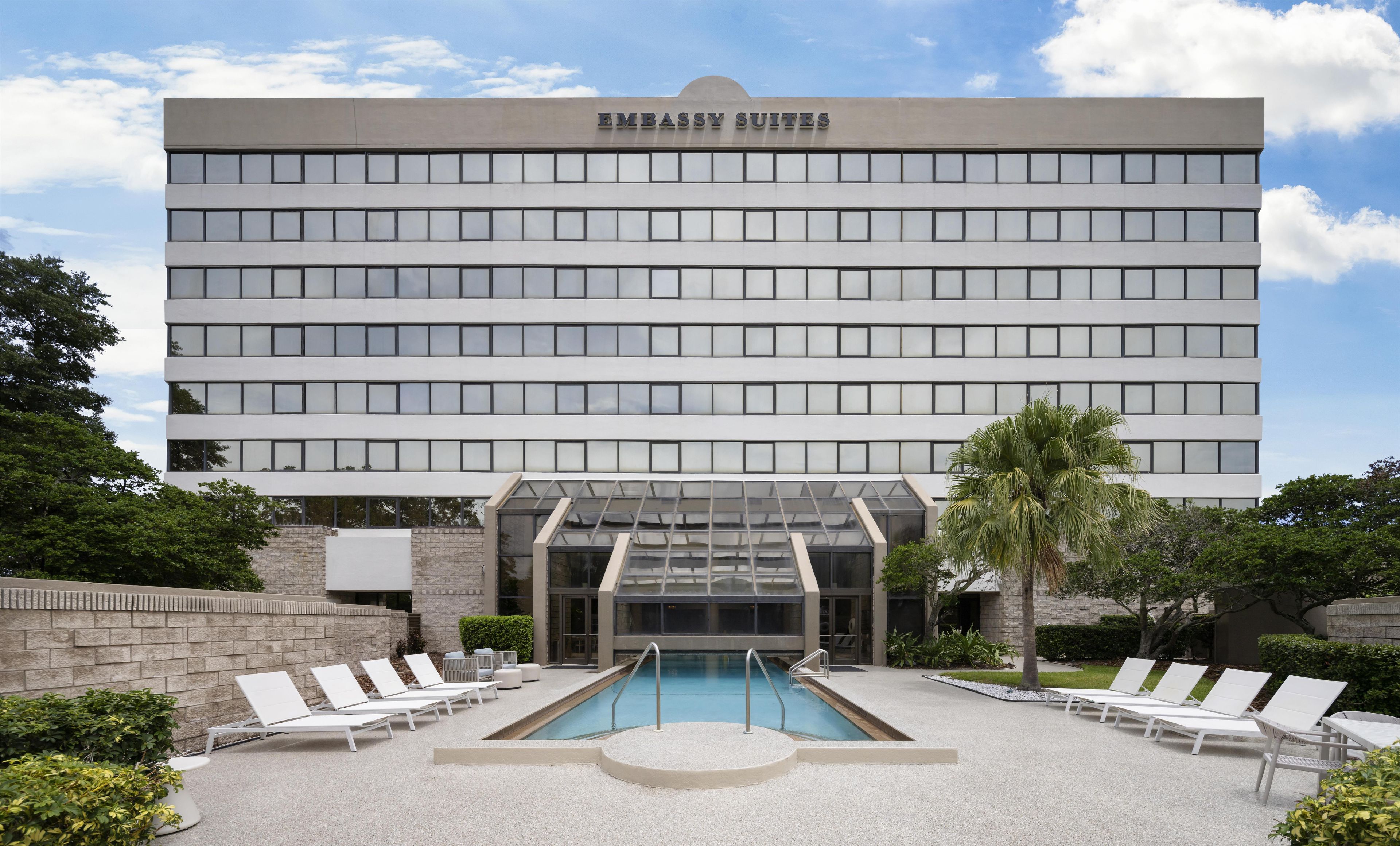 A building with 'EMBASSY SUITES' signage, a swimming pool, and lounge chairs.