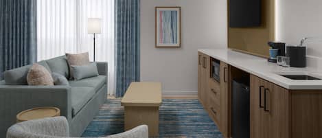 A hotel room with a grey sofa, a wooden cabinet with a sink, a small table, and a blue patterned carpet.