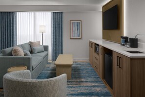 Une chambre d’hôtel avec un canapé gris, un meuble en bois avec un lavabo, une petite table et un tapis bleu à motifs.