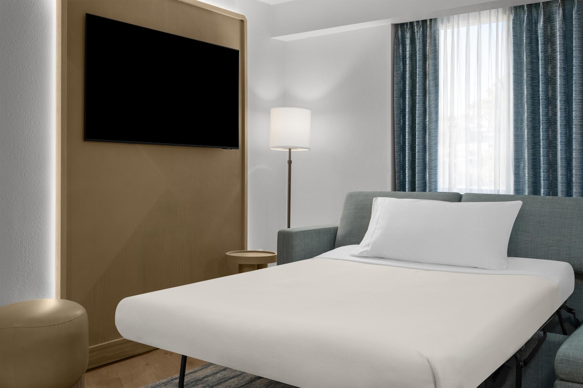 A hotel room with a bed, a TV mounted on the wall, a lamp, and a window with curtains.