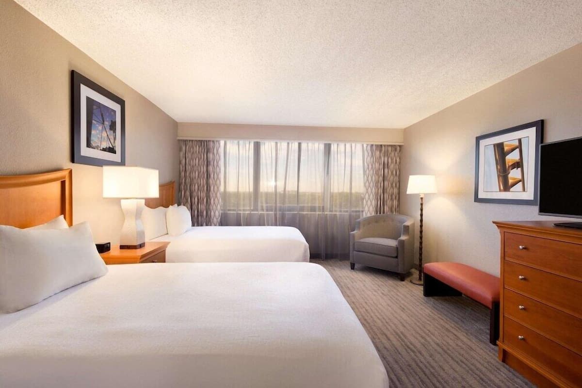 A hotel room with two beds, a desk, a chair, a TV, and a window with curtains.