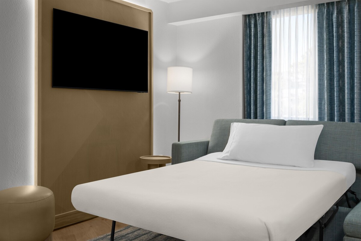 A hotel room with a bed, a TV mounted on the wall, a lamp, and a window with curtains.