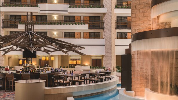 A multi-level hotel lobby with a central pool, a bar area, and a seating area with tables and chairs.