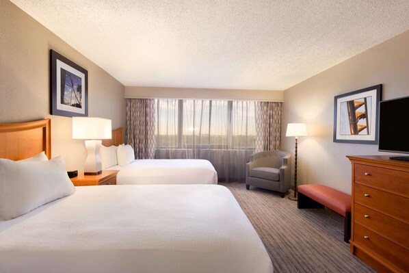 Two large beds with white linens and a wooden headboard, a dresser, flat-screen TV, and a chair by the window.