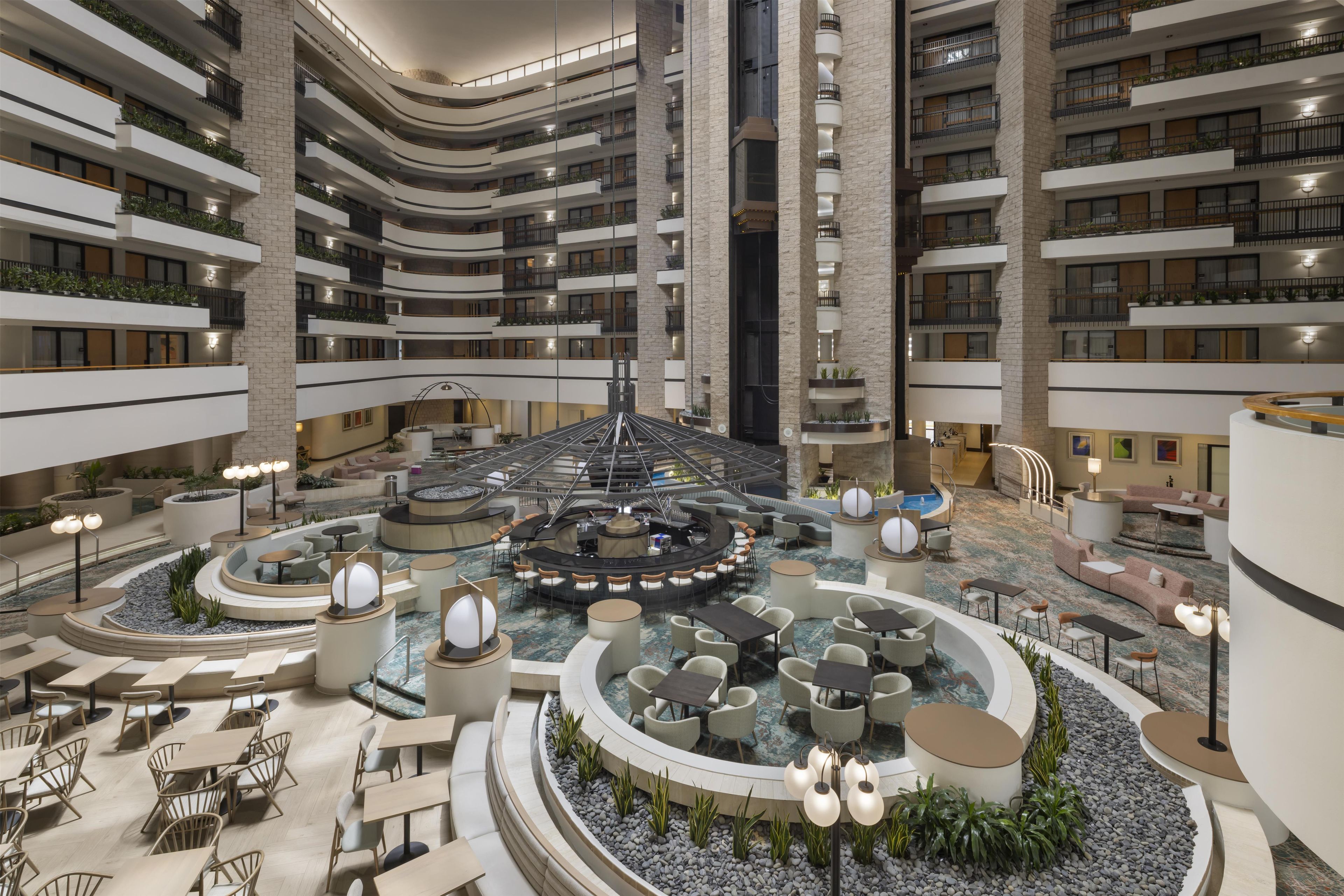 A multi-level hotel atrium with a central bar area, seating arrangements, and a pool.