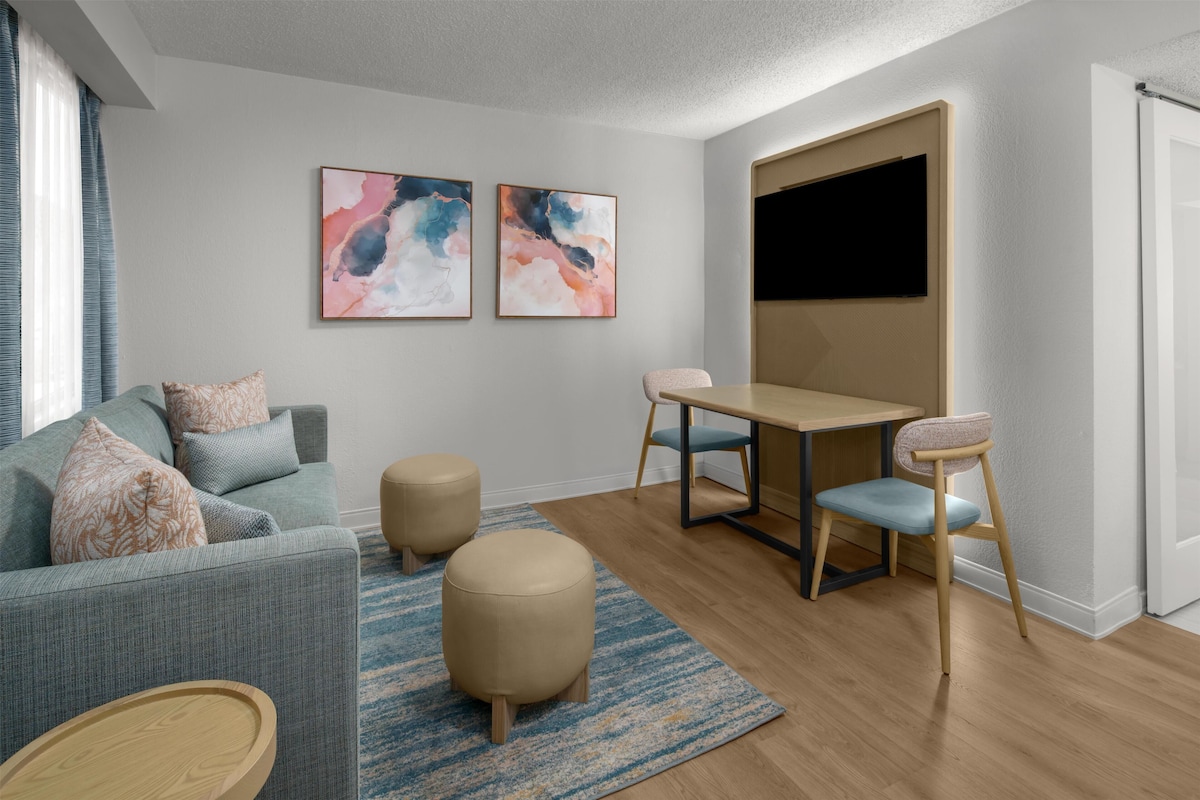 Una sala de estar moderna con un sofá, otomanas, una mesa y sillas. Hay pinturas abstractas en la pared y un televisor de pantalla plana montado sobre la mesa.