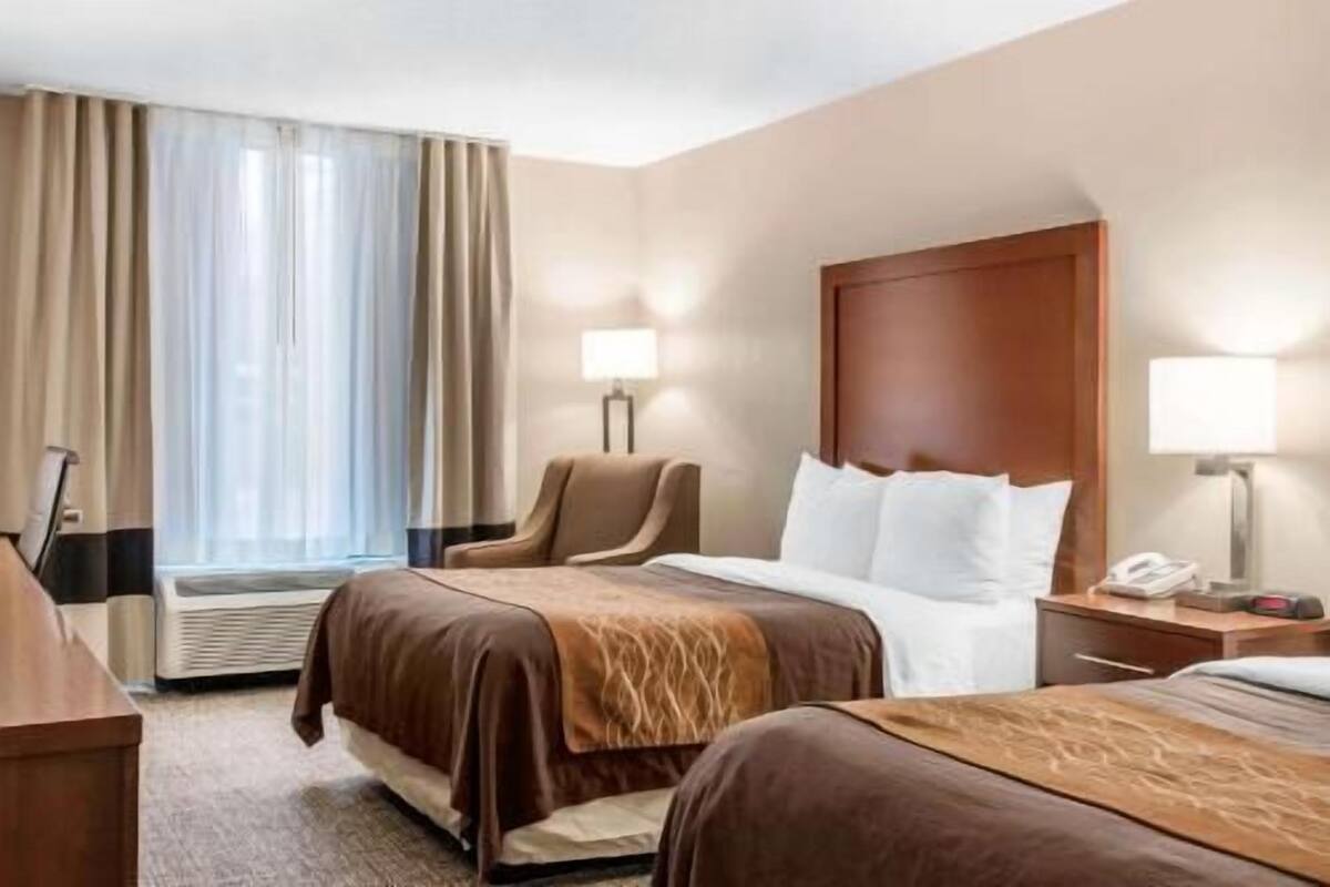 A hotel room with two beds, a chair, a desk, a lamp, and a window with curtains.