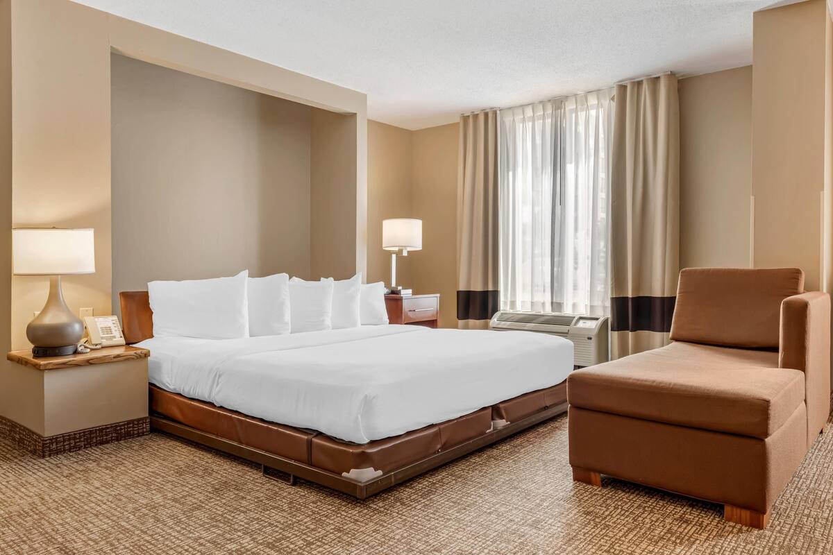 A hotel room with a large bed, a chair, a nightstand with a lamp, and a window with curtains.