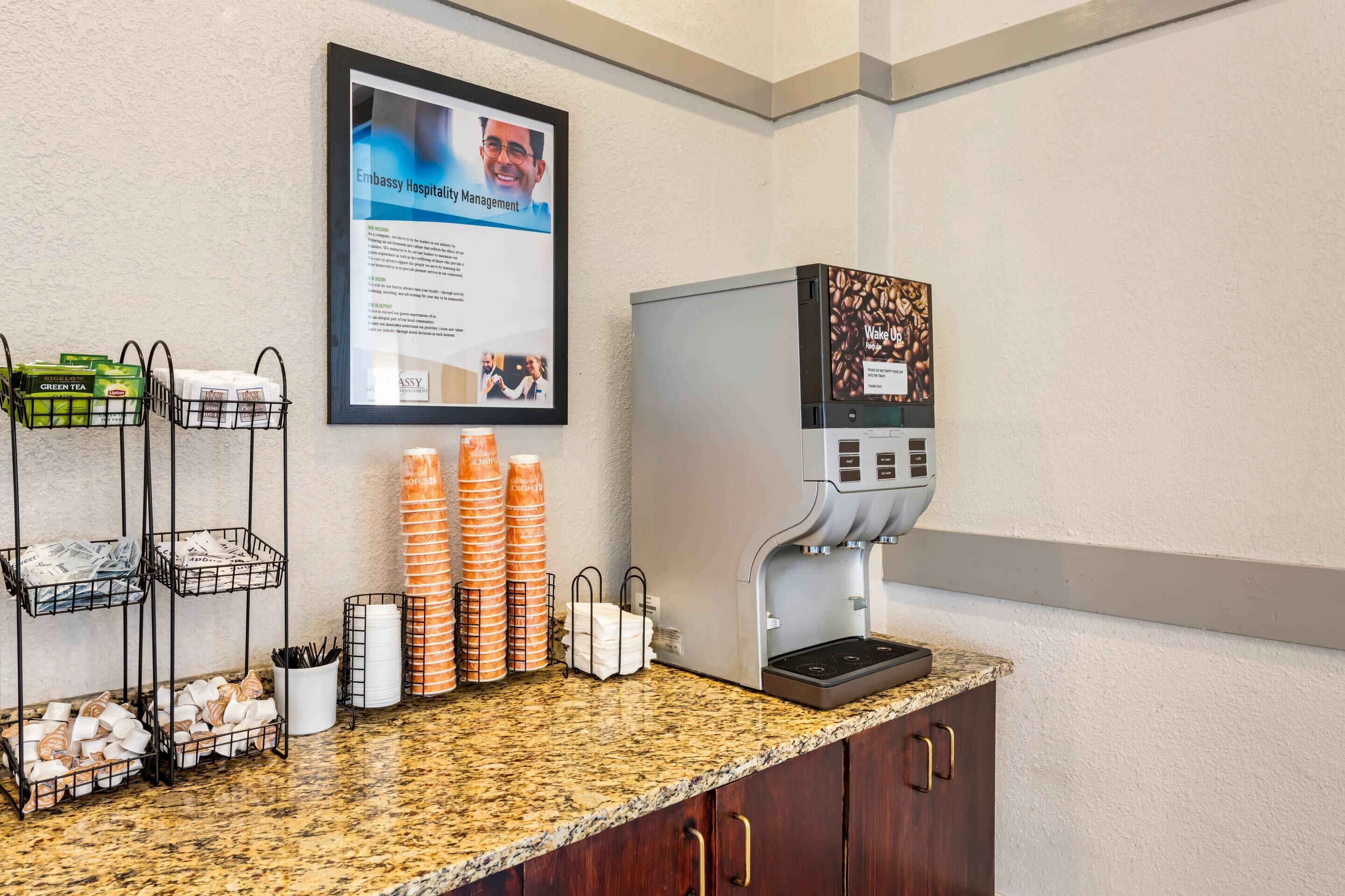 A coffee station with a coffee machine, paper cups, and a sign about embassy hospitality management.