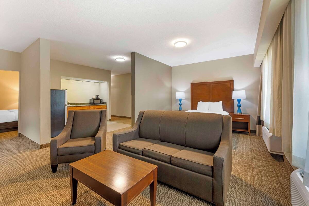 Habitación de hotel con sofá, sillón y una mesita. Posee una cama con cabecero de madera y dos mesitas de noche con lámparas.