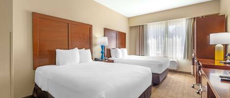 A hotel room with two large beds, a desk with a chair, a lamp, and a window with curtains.