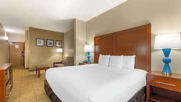 A hotel room with a large bed, two bedside tables with lamps, a wooden headboard, a small desk with a chair, and a door leading to a bathroom.