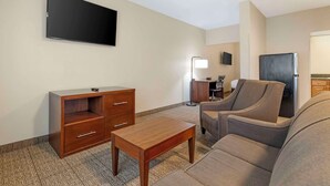 Habitación de hotel con televisor de pantalla plana montado en la pared, un mueble de madera con cajones, una pequeña mesa de madera, un sofá gris y una lámpara de pie.