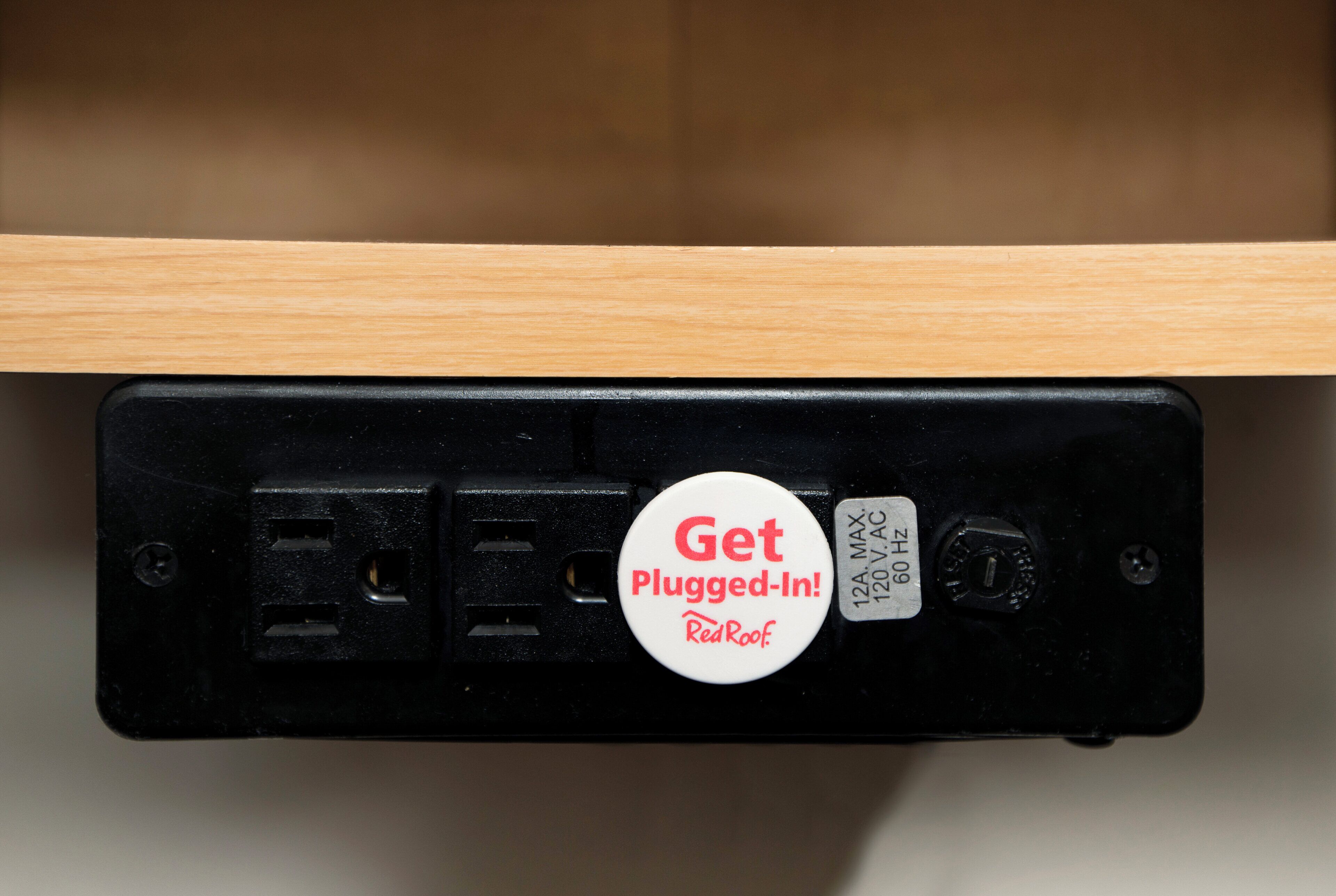 Un enchufe eléctrico negro con un dispositivo blanco insertado, etiquetado 'Get Plugged-In! RedRoof', y una placa metálica con especificaciones eléctricas.