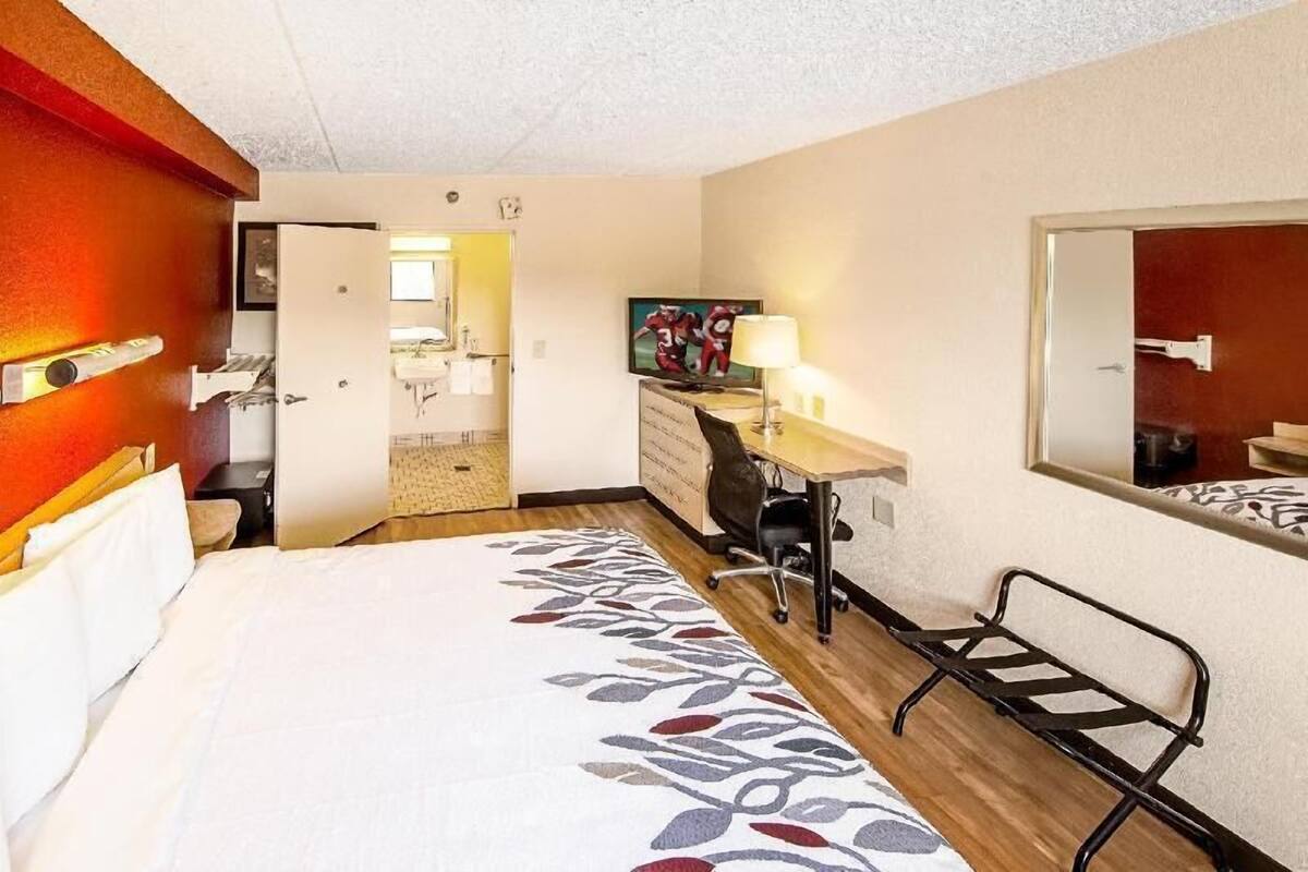 A hotel room with a bed, desk, chair, and a mirror. There is a door leading to a bathroom and a hallway.