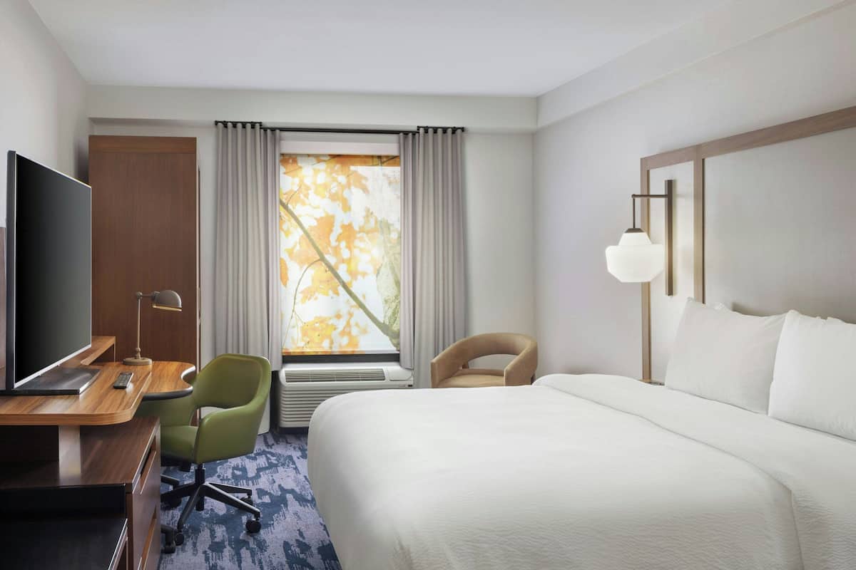 Ein Hotelzimmer mit einem Bett, einem Schreibtisch mit Fernseher, einem Stuhl, einem Fenster mit Vorhängen und einer Wandlampe.